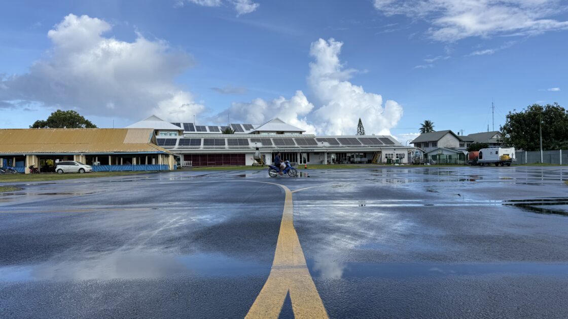 朝の雨上がり、濡れた滑走路にツバルの青空と雲、空港の建物が反射している様子。