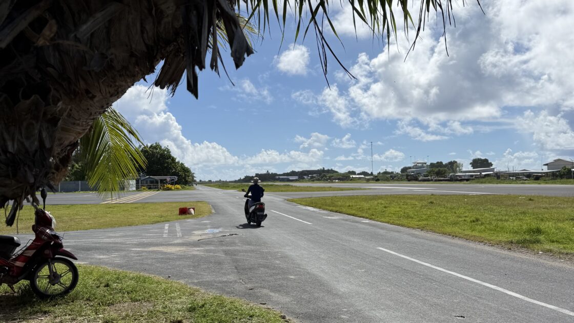 青空と雲の下、ツバルの滑走路横の道路でバイクの慣らし運転をしている様子。