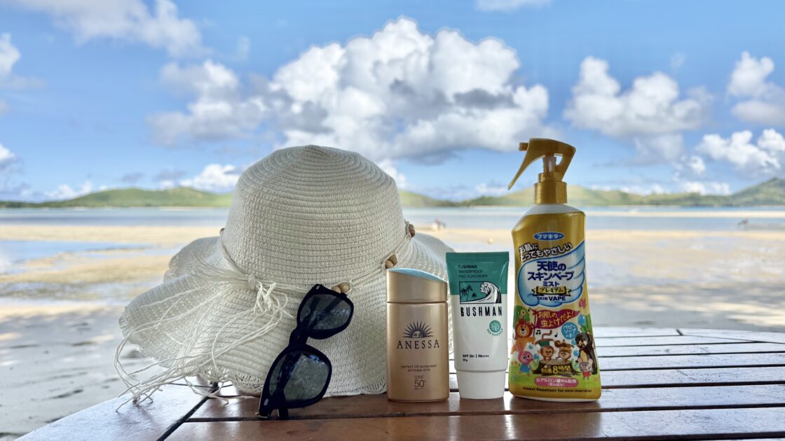 ツバルの青空と海を背景に並べた麦わら帽子、UNDERGROUNDJOBSの偏光サングラス、BUSHMANの日焼け止め、スキンベープ天使のミストプレミアム